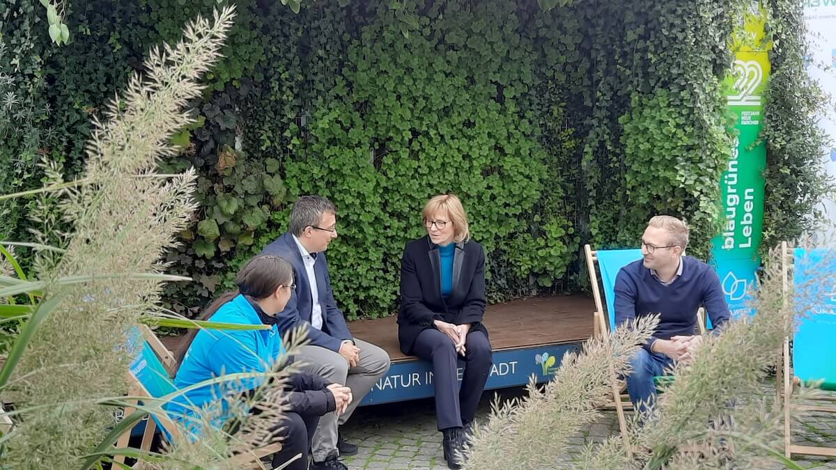 Vier Personen sitzen auf einer Holzbühne und Liegestühlen in Gelsenkirchen und unterhalten sich. Im Hintergrund ist eine Klimawerk-Stele zu sehen.
