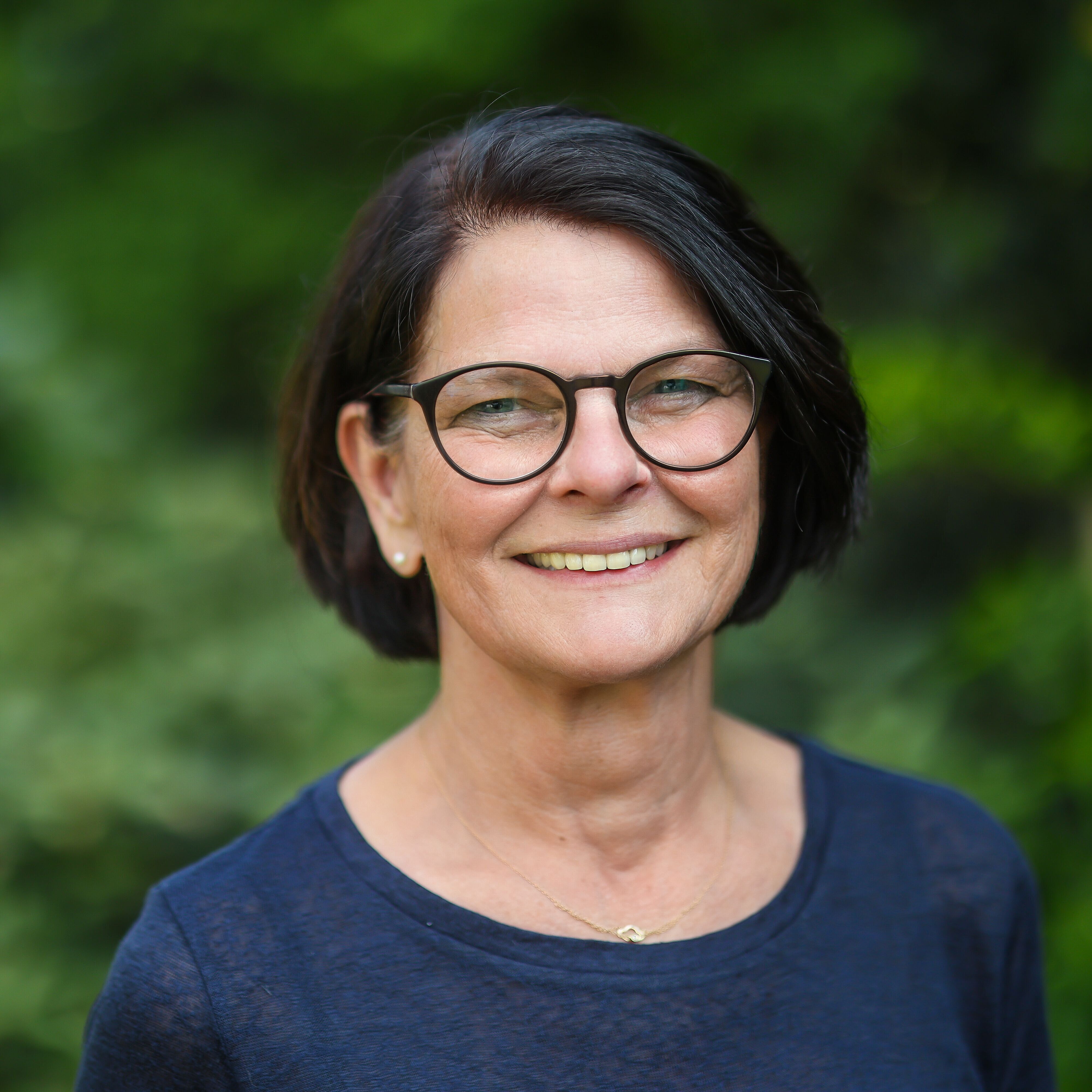 Das Portrait einer Frau mit braunen Haaren und blauem Shirt. Sie hat helle Haut und eine Brille auf.